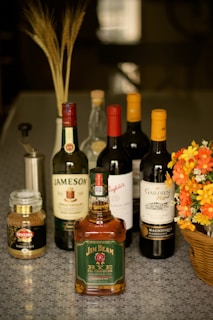 A selection of liquor bottles, including whiskey and wine. They are arranged neatly on a speckled gray surface alongside a jar of instant coffee and a wicker basket holding vibrant orange and yellow flowers. In the background, there are decorative wheat stalks in a holder.