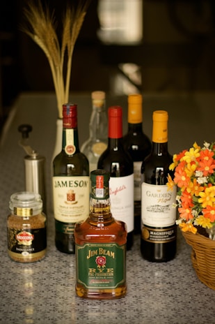 A selection of liquor bottles, including whiskey and wine. They are arranged neatly on a speckled gray surface alongside a jar of instant coffee and a wicker basket holding vibrant orange and yellow flowers. In the background, there are decorative wheat stalks in a holder.