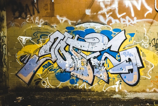 A vibrant graffiti mural on a brick wall featuring bold, abstract shapes and layers of paint in various colors. The central graffiti art is primarily in blue, yellow, and white hues, with surrounding tags and smaller pieces in different colors. The background shows a worn brick texture with additional tags and markings.