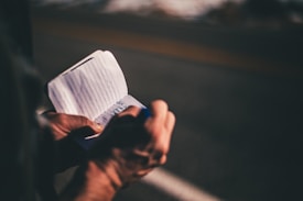 A person is holding a small notebook and writing on it with a pen. The notebook has lined pages and the background appears to be a road or outdoor setting with a blurred effect.