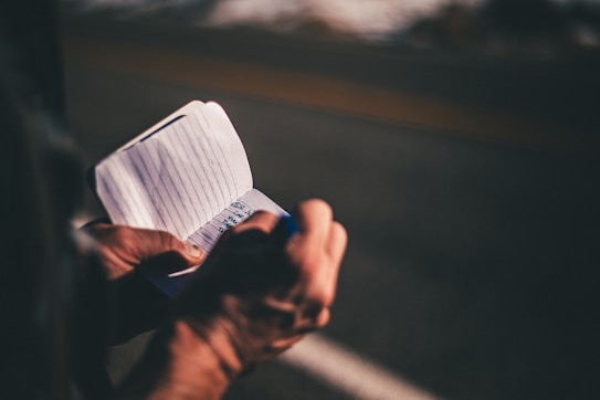 A person is holding a small notebook and writing on it with a pen. The notebook has lined pages and the background appears to be a road or outdoor setting with a blurred effect.