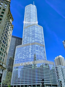 A tall, modern skyscraper with a reflective glass facade. The structure is composed of several stacked cylindrical tiers, tapering off at the top, with a large sign featuring capital letters. Surrounding buildings are visible, including one with a classical architectural style.