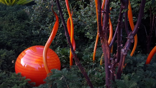 Orange and red glass sculptures resembling plant forms are placed among lush green foliage. A large, spherical object with orange stripes is nestled in the greenery, accompanied by several tall, curling shapes that mimic stems or flames. The scene evokes an artistic blend of nature and sculpture.