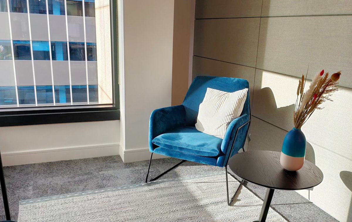 Sillón de terciopelo azul frío junto a una ventana, con pared beige cálida, alfombra gris y un jarrón con pampas