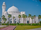A large white mosque with a prominent dome and minaret is surrounded by palm trees and a well-maintained lawn. The sky is bright and clear, adding to the serene ambiance. A red jogging track runs along the front, lined with lampposts and signs.