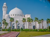 A large white mosque with a prominent dome and minaret is surrounded by palm trees and a well-maintained lawn. The sky is bright and clear, adding to the serene ambiance. A red jogging track runs along the front, lined with lampposts and signs.