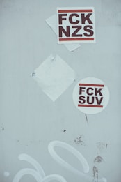 Two stickers with bold text, one square and one circular, are displayed on a light gray surface. The square sticker has the letters 'FCK NZS' in a strong font, while the round sticker reads 'FCK SUV'. A blank, white paper backing is also partially visible underneath one of the stickers.