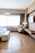 A hotel room with a modern design features a large window covered with sheer curtains, allowing natural light to fill the space. A comfortable bed with a white cover and a cushioned bench at the foot is placed to the left. A plush armchair sits in the corner next to a wooden desk, decorated with a lamp and some accessories. A flat-screen TV is mounted on the wall opposite the bed. The flooring is wooden, adding warmth to the neutral color palette of the room.