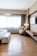 A hotel room with a modern design features a large window covered with sheer curtains, allowing natural light to fill the space. A comfortable bed with a white cover and a cushioned bench at the foot is placed to the left. A plush armchair sits in the corner next to a wooden desk, decorated with a lamp and some accessories. A flat-screen TV is mounted on the wall opposite the bed. The flooring is wooden, adding warmth to the neutral color palette of the room.