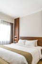 A neatly made bed with white linens and pillows, featuring a decorative runner, is positioned against a wooden headboard. A stylish wall lamp is mounted on the wooden panel beside the bed. Natural light filters through a sheer curtain covering a large window, casting a soft glow into the room.