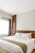 A neatly made bed with white linens and pillows, featuring a decorative runner, is positioned against a wooden headboard. A stylish wall lamp is mounted on the wooden panel beside the bed. Natural light filters through a sheer curtain covering a large window, casting a soft glow into the room.