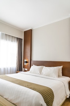 A neatly made bed with white linens and pillows, featuring a decorative runner, is positioned against a wooden headboard. A stylish wall lamp is mounted on the wooden panel beside the bed. Natural light filters through a sheer curtain covering a large window, casting a soft glow into the room.