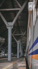 A train station platform with a long metal roof supported by large beams. A train is on the right side with blue and orange stripes. A person stands on the platform, looking at a mobile device, surrounded by industrial architecture.