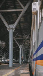 A train station platform with a long metal roof supported by large beams. A train is on the right side with blue and orange stripes. A person stands on the platform, looking at a mobile device, surrounded by industrial architecture.