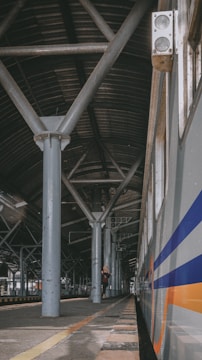 A train station platform with a long metal roof supported by large beams. A train is on the right side with blue and orange stripes. A person stands on the platform, looking at a mobile device, surrounded by industrial architecture.
