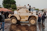 An outdoor scene with a tan armored vehicle displayed on wet cobblestones. Several people, including a child, are interacting with the vehicle, some climbing on it. There are others walking around, with tents and trees visible in the background and a few buildings further away.
