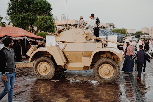 An outdoor scene with a tan armored vehicle displayed on wet cobblestones. Several people, including a child, are interacting with the vehicle, some climbing on it. There are others walking around, with tents and trees visible in the background and a few buildings further away.