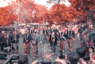 A cultural procession with people dressed in traditional attire walking through a street lined with orange-hued trees. The participants are wearing patterned sarongs and matching tops, and some are holding torches. The scene is lively with onlookers capturing the event using cameras.