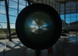 A large cylindrical object is centrally positioned, possibly the engine or exhaust of a jet or aircraft, with light reflecting off its inner surface. The setting is indoors, with large windows allowing natural light to illuminate the space. In the background, part of an aircraft fuselage is visible.