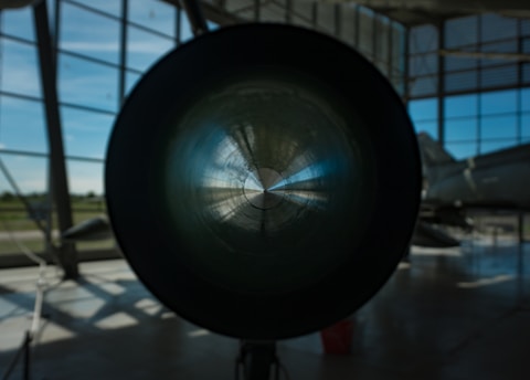 A large cylindrical object is centrally positioned, possibly the engine or exhaust of a jet or aircraft, with light reflecting off its inner surface. The setting is indoors, with large windows allowing natural light to illuminate the space. In the background, part of an aircraft fuselage is visible.