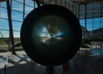 A large cylindrical object is centrally positioned, possibly the engine or exhaust of a jet or aircraft, with light reflecting off its inner surface. The setting is indoors, with large windows allowing natural light to illuminate the space. In the background, part of an aircraft fuselage is visible.
