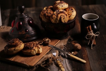 A wooden table is set with a rustic scene featuring freshly baked cinnamon rolls drizzled with chocolate on a wooden cutting board and in a dark ceramic bowl. Beside them lies a vintage knife, whole walnuts, and a black mug filled with coffee. A bundle of cinnamon sticks tied with twine rests next to the mug. The background elements include a metal decorative piece with red accents and a burlap cloth.
