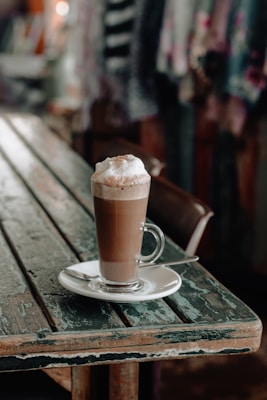A latte with frothy milk served in a tall glass cup sits on a white saucer with a silver spoon on an aged wooden table. The background features blurred colorful fabrics, suggesting an artistic or cozy environment.