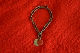 A metal chain with two round tags rests on a textured red fabric. The fabric has a quilted pattern with a diamond shape and leaf motifs. The chain and tags are positioned in a circular form.
