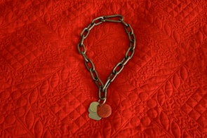 A metal chain with two round tags rests on a textured red fabric. The fabric has a quilted pattern with a diamond shape and leaf motifs. The chain and tags are positioned in a circular form.