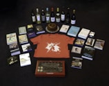 A collection of items is arranged on a dark floor, including a variety of books, several bottles of wine, a red t-shirt with a graphic design, a round cake on a silver platter, and a box of chocolates. There are also some DVDs and CDs, each with distinct cover art.