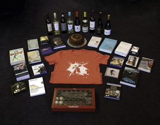 A collection of items is arranged on a dark floor, including a variety of books, several bottles of wine, a red t-shirt with a graphic design, a round cake on a silver platter, and a box of chocolates. There are also some DVDs and CDs, each with distinct cover art.
