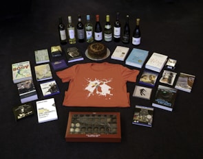 A collection of items is arranged on a dark floor, including a variety of books, several bottles of wine, a red t-shirt with a graphic design, a round cake on a silver platter, and a box of chocolates. There are also some DVDs and CDs, each with distinct cover art.