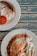 A rustic wooden table serves as the backdrop for two plates of breakfast food. One plate contains a sausage, a sunny-side-up egg, and a slice of grilled tomato. The other plate features an omelet with a side of sliced cucumber and lettuce garnish.