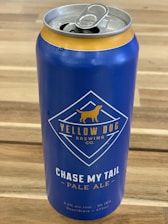 A blue aluminum can of beer with a pull-tab lid, featuring the logo and branding of Yellow Dog Brewing Co. The label includes the text 'Chase My Tail Pale Ale' and details like the alcohol percentage (5.2% alc/vol) and volume (473ml). The can is placed on a wooden surface.