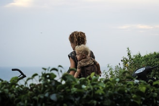 A woman with curly hair is carrying a young child on her back, surrounded by lush green foliage with a view of the ocean in the background. A motorcycle's handlebars are partially visible indicating a journey or travel scenario.