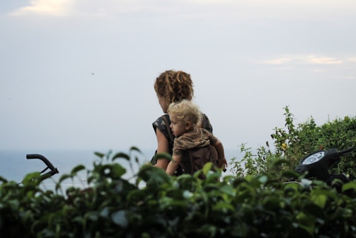 A woman with curly hair is carrying a young child on her back, surrounded by lush green foliage with a view of the ocean in the background. A motorcycle's handlebars are partially visible indicating a journey or travel scenario.