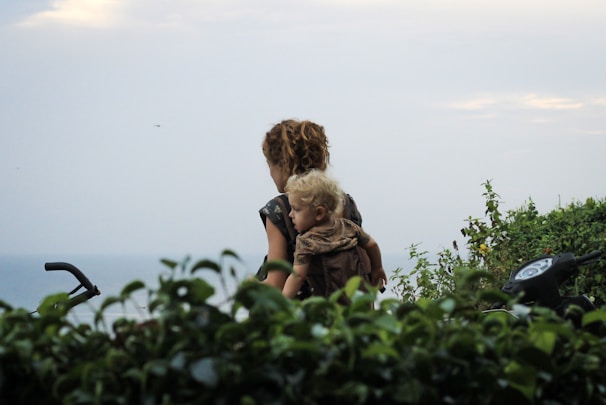 A woman with curly hair is carrying a young child on her back, surrounded by lush green foliage with a view of the ocean in the background. A motorcycle's handlebars are partially visible indicating a journey or travel scenario.