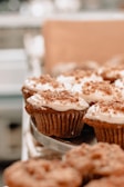 A close-up view of several frosted cupcakes with a crumb topping arranged on a tray. The background is blurred, suggesting a bakery setting or kitchen environment.