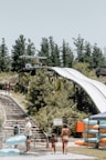A water park scene with people preparing to use a large slide. There are tall trees in the background, a set of wooden stairs on the left, and stacks of inflatable rings on the right. A canopy tent is visible with more rings underneath. The slide structure features a platform with a roof, surrounded by lush green vegetation.