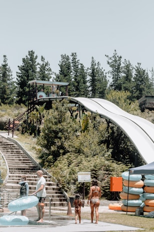 A water park scene with people preparing to use a large slide. There are tall trees in the background, a set of wooden stairs on the left, and stacks of inflatable rings on the right. A canopy tent is visible with more rings underneath. The slide structure features a platform with a roof, surrounded by lush green vegetation.