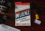A book titled 'Influence: The Psychology of Persuasion' by Robert B. Cialdini is under a pair of sunglasses on a reddish-brown wooden surface. A camera with visible lens details is placed on the left. There is a roll of Kodak Gold 200 film and a small white container nearby.