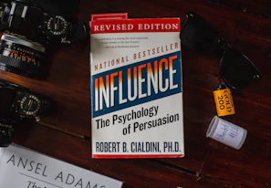A book titled 'Influence: The Psychology of Persuasion' by Robert B. Cialdini is under a pair of sunglasses on a reddish-brown wooden surface. A camera with visible lens details is placed on the left. There is a roll of Kodak Gold 200 film and a small white container nearby.
