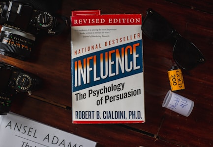 A book titled 'Influence: The Psychology of Persuasion' by Robert B. Cialdini is under a pair of sunglasses on a reddish-brown wooden surface. A camera with visible lens details is placed on the left. There is a roll of Kodak Gold 200 film and a small white container nearby.