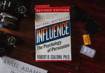 A book titled 'Influence: The Psychology of Persuasion' by Robert B. Cialdini is under a pair of sunglasses on a reddish-brown wooden surface. A camera with visible lens details is placed on the left. There is a roll of Kodak Gold 200 film and a small white container nearby.