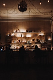 A dimly lit bar with a variety of bottles arranged on shelves behind the counter. Three silhouetted individuals sit at the bar, which is decorated with soft, warm lighting. An ornate ceiling with a reflective disco ball and exposed bulb lights adds to the ambiance.