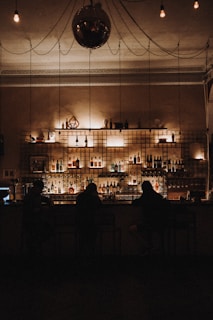A dimly lit bar with a variety of bottles arranged on shelves behind the counter. Three silhouetted individuals sit at the bar, which is decorated with soft, warm lighting. An ornate ceiling with a reflective disco ball and exposed bulb lights adds to the ambiance.