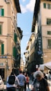 A busy street scene with historical European architecture featuring tall buildings with green shutters. Several people are walking in the street, some with backpacks. Signage includes 'Hotel' and 'Palazzo Benci'.