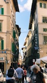 A busy street scene with historical European architecture featuring tall buildings with green shutters. Several people are walking in the street, some with backpacks. Signage includes 'Hotel' and 'Palazzo Benci'.
