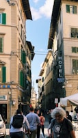 A busy street scene with historical European architecture featuring tall buildings with green shutters. Several people are walking in the street, some with backpacks. Signage includes 'Hotel' and 'Palazzo Benci'.