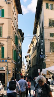 A busy street scene with historical European architecture featuring tall buildings with green shutters. Several people are walking in the street, some with backpacks. Signage includes 'Hotel' and 'Palazzo Benci'.